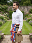 A man standing in a garden wearing a white dress shirt, black bow tie, and a tan kilt with a Union Jack flag design on the front panel.