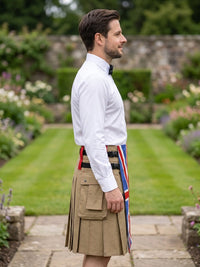 A man standing in profile in a garden wearing a white long-sleeve dress shirt, a tan pleated kilt, a black bow tie, and a red, white, and blue sash.