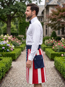 A man in a white dress shirt and black bow tie stands in profile in a garden setting, wearing a traditional Scottish kilt with red and white vertical stripes, a navy blue waist belt, and a matching navy blue front pouch pocket.