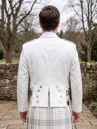 A man viewed from behind wearing a white formal jacket over a white and gray plaid kilt, standing outdoors on a stone path with trees and a stone wall in the background.