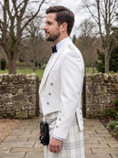 A man in profile wearing a white double-breasted formal jacket, a white plaid kilt, a black bow tie, and a black sporran, standing outdoors on a stone path with trees and a stone wall in the background.