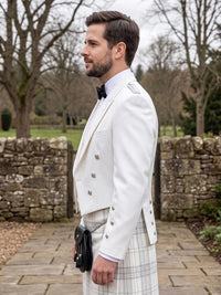 A man in profile wearing a white double-breasted formal jacket, a white plaid kilt, a black bow tie, and a black sporran, standing outdoors on a stone path with trees and a stone wall in the background.