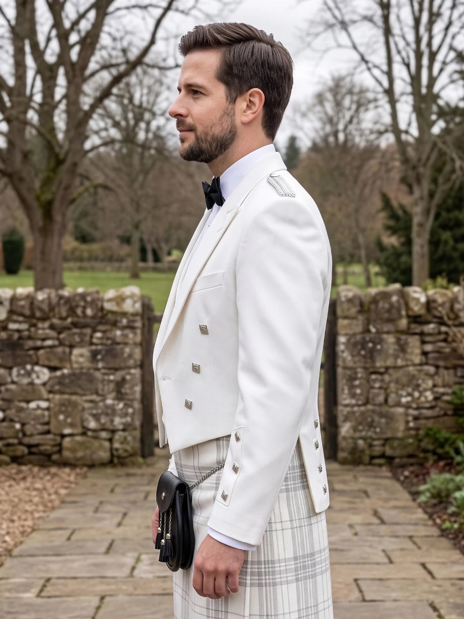 A man in profile wearing a white double-breasted formal jacket, a white plaid kilt, a black bow tie, and a black sporran, standing outdoors on a stone path with trees and a stone wall in the background.