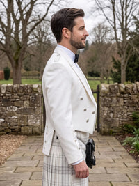 A man in profile wearing a white double-breasted formal jacket with silver buttons, a white shirt, a black bow tie, and a white plaid kilt, standing outdoors on a stone path with a stone wall and bare trees in the background.