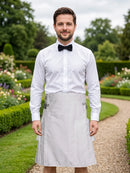 A man standing in a garden wearing a white long-sleeve dress shirt, black bow tie, and a light gray pleated kitchen apron tied around his waist.
