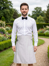 A man standing in a garden wearing a white long-sleeve dress shirt, black bow tie, and a light gray pleated kitchen apron tied around his waist.