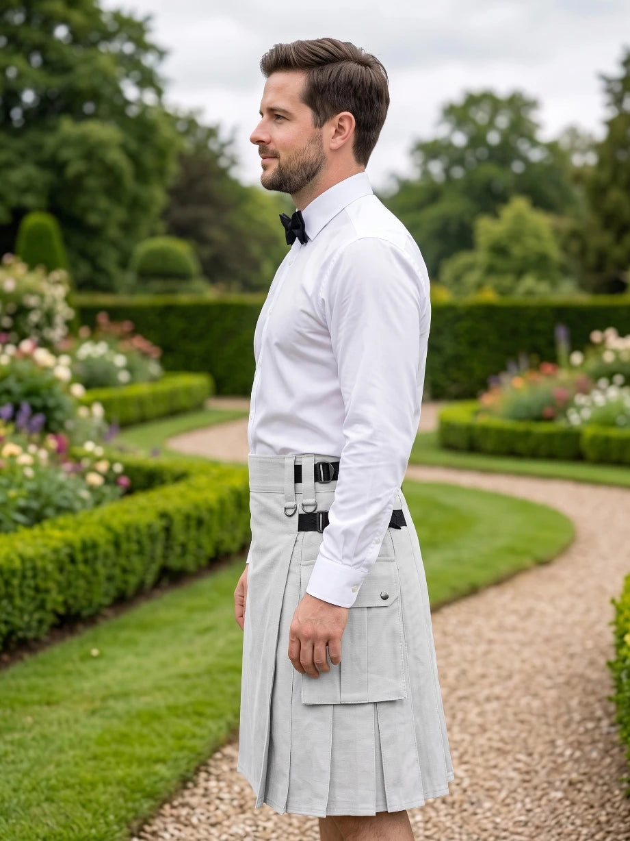 A man standing in profile in a formal garden, wearing a white long-sleeve dress shirt, black bow tie, and a light gray pleated kilt with black belt details.
