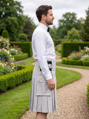 A man in a white dress shirt and black bow tie stands in profile outdoors wearing a light gray pleated kilt with flap pockets and a black belt, set against a manicured garden path and hedges.