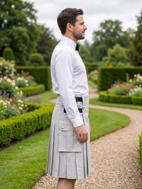 A man in a white dress shirt and black bow tie stands in profile outdoors wearing a light gray pleated kilt with flap pockets and a black belt, set against a manicured garden path and hedges.