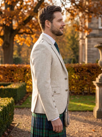A man in profile wearing a light beige tweed blazer, a green and black tartan kilt, a white shirt, and a dark green tie, standing outdoors in a garden with autumn trees and hedges.
