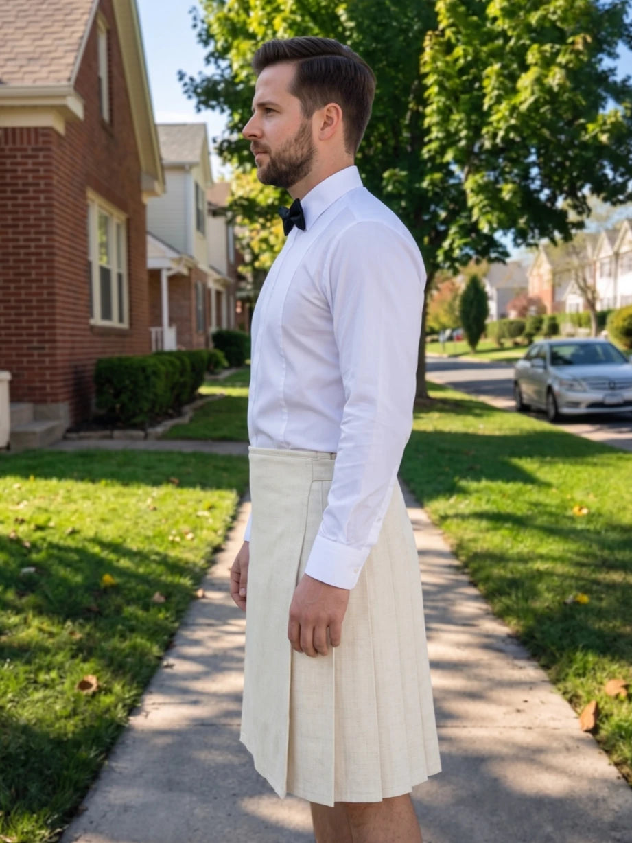 A man wearing a white dress shirt, black bow tie, and a pleated cream-colored kilt stands on a suburban sidewalk.