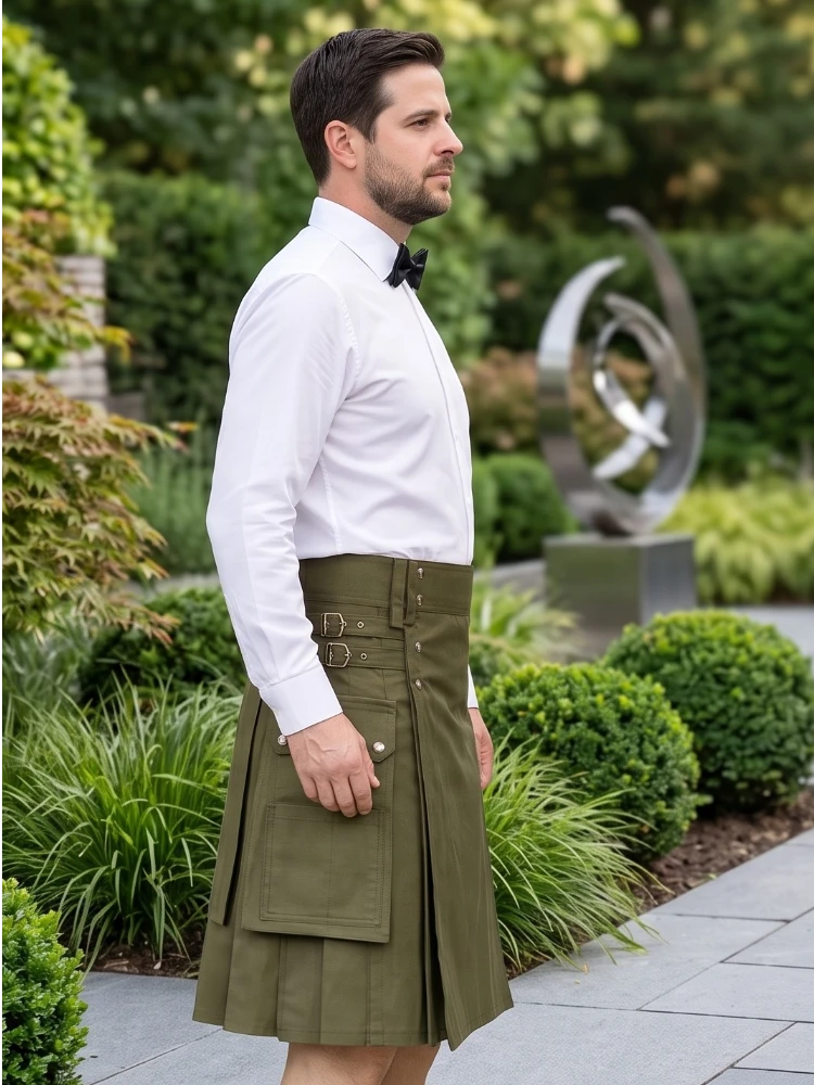 A man wearing a white dress shirt, black bow tie, and an olive green kilt with belt details, standing in a landscaped garden setting.