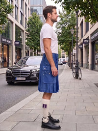 A man standing on a city sidewalk wearing a blue denim kilt, white t-shirt, white socks, and black leather shoes, with a street scene and parked cars in the background.