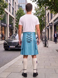 A man standing on a city sidewalk, photographed from behind, wearing a light blue pleated kilt with black lace-up detailing at the back, a white t-shirt, white knee-high socks, and black shoes. Buildings, trees, a black Mercedes, and a bicycle are visible in the background.