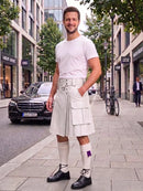 A man stands on a city sidewalk wearing a white short-sleeve t-shirt and a pleated white kilt-style skirt with a matching belt, paired with white knee-high socks and black lace-up shoes. Buildings, trees, a parked black car, and a bicycle are visible in the urban street setting.