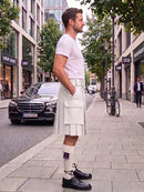 A man wearing a white short-sleeve t-shirt and a white pleated kilt with a white belt, standing on a city sidewalk next to a street with a black Mercedes-Benz car and trees.