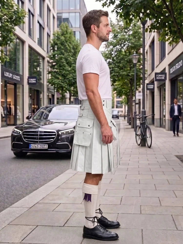 A man standing on a city sidewalk wearing a white short-sleeve T-shirt and a light gray pleated kilt with white socks and black shoes. Buildings, trees, a black Mercedes-Benz car, and a bicycle are visible in the urban street setting.